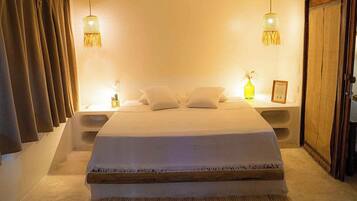 Cama bem arrumada com colcha e travesseiros brancos, duas luminárias de cabeceira, mesa de cabeceira e guarda-roupa de madeira.