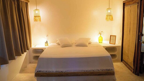 A neatly made bed with a white bedspread and pillows, two bedside lamps, a nightstand, and a wooden wardrobe. - Aloha Village Hotel (Amontada)