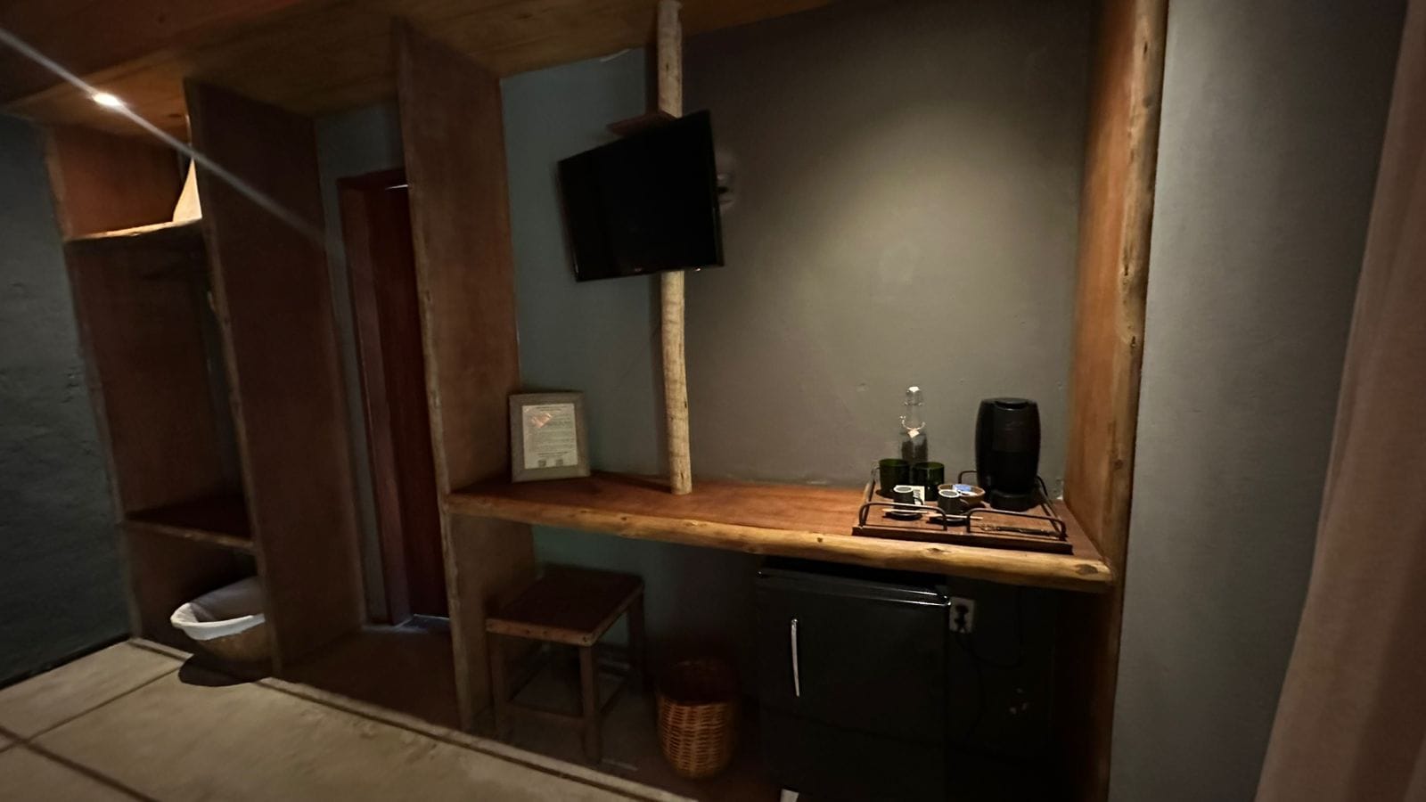 Cocina compacta con encimera de madera, un taburete pequeño, una cafetera y un televisor fijado a la pared.