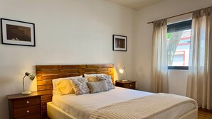 A bedroom with a wooden headboard, a large bed with white bedding, two bedside lamps, a small nightstand, a framed picture on the wall, and a window with curtains. - BELLA CENA by Azores HomeMade (Ponta Delgada)