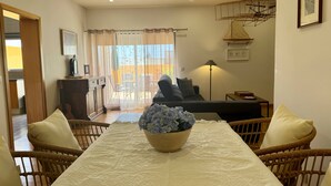 A dining area with a table set for two, a wicker chair, a sofa, and a lamp. - BELLA CENA by Azores HomeMade (Ponta Delgada)
