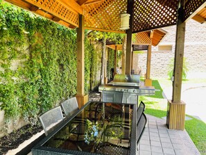 Een terras buiten met een tafel met glazen blad, metalen stoelen en een roestvrijstalen barbecue.