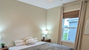 Un dormitorio con cama, mesita de noche, lámpara, ventilador de techo, ventana con cortinas y alfombra estampada.