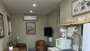 Una kitchenette compacta con pileta, estufa y microondas, un pequeño comedor con mesa y sillas, y un televisor colocado en la pared.