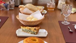 Una mesa de comedor dispuesta con pan, una quiche, un pastel y un centro de mesa floral.