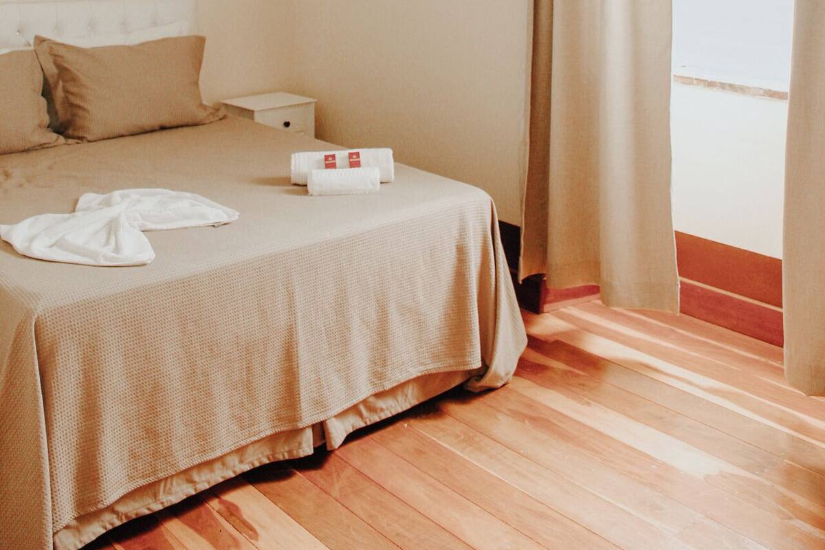 A hotel room with a bed, a nightstand, a chandelier, and a window with curtains.