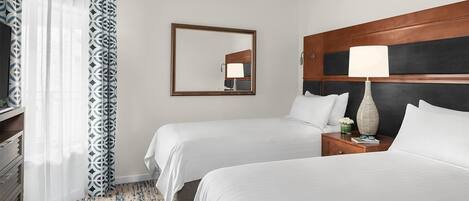 A hotel room with two beds, a wooden headboard, a nightstand with a lamp, a mirror, and a patterned curtain.