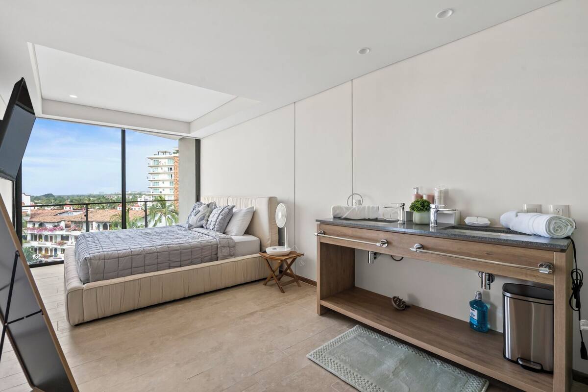 Une chambre d’hôtel moderne avec un grand lit, une coiffeuse en bois, une petite table d’appoint et une vue sur la ville.