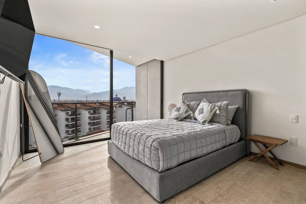 Une chambre à coucher avec un lit, des oreillers, une table de chevet et une vue sur des immeubles et des montagnes.