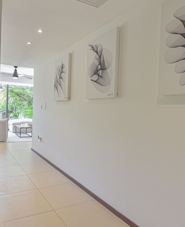 A hallway with two framed artworks on the wall, a ceiling fan, and a view of an outdoor area with greenery.
