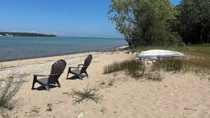 En strand med två solstolar och ett avskärmat område, omgiven av träd och med fri utsikt över vattnet.