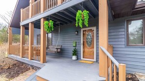 A two-story house with a porch, wooden railings, and hanging green plants.