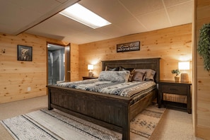 A wooden-paneled bedroom with a bed, two bedside tables, a wall-mounted sign, and a door. - Brown Bear Cabin - 3BR w/ Hot Tub and Lake Access (Nekoosa)