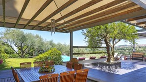 A covered outdoor dining area with a wooden table and chairs, surrounded by a garden and a view of a swimming pool.