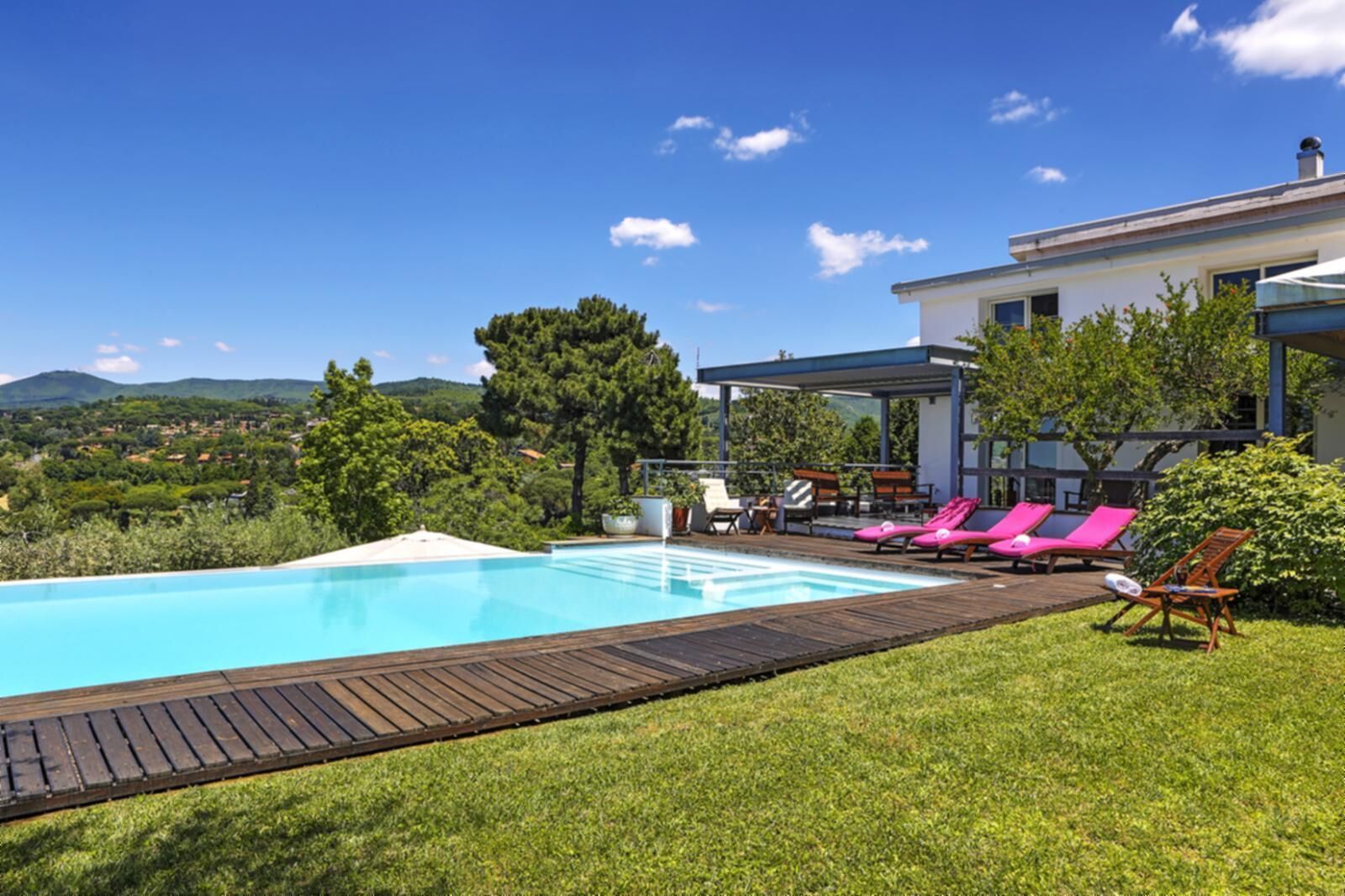 Piscina con plataforma de madera, rodeada de césped y árboles, con vista a un paisaje montañoso.