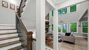 Una escalera de madera con barandillas negras que conduce a una sala de estar con chimenea, un sofá y grandes ventanales con vistas a zonas verdes.