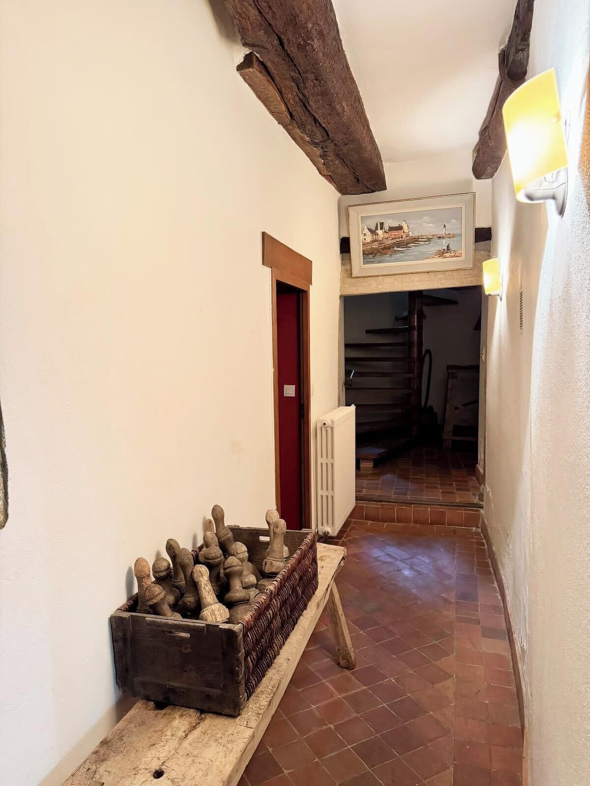 Un couloir étroit avec des poutres en bois rustiques, un sol carrelé et un coffre en bois rempli de petits objets.