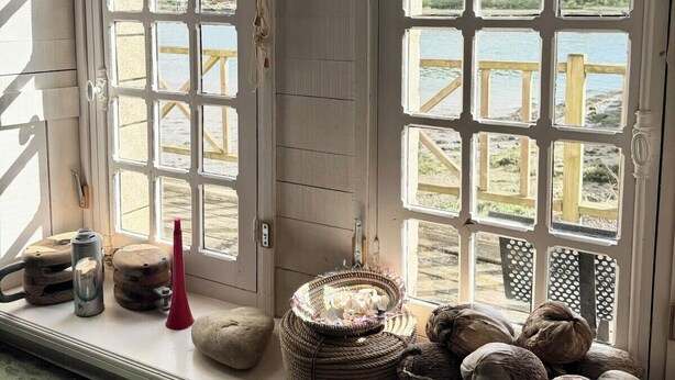 A rustic room with large windows, a wooden bench, a clock, and various items on the windowsill.