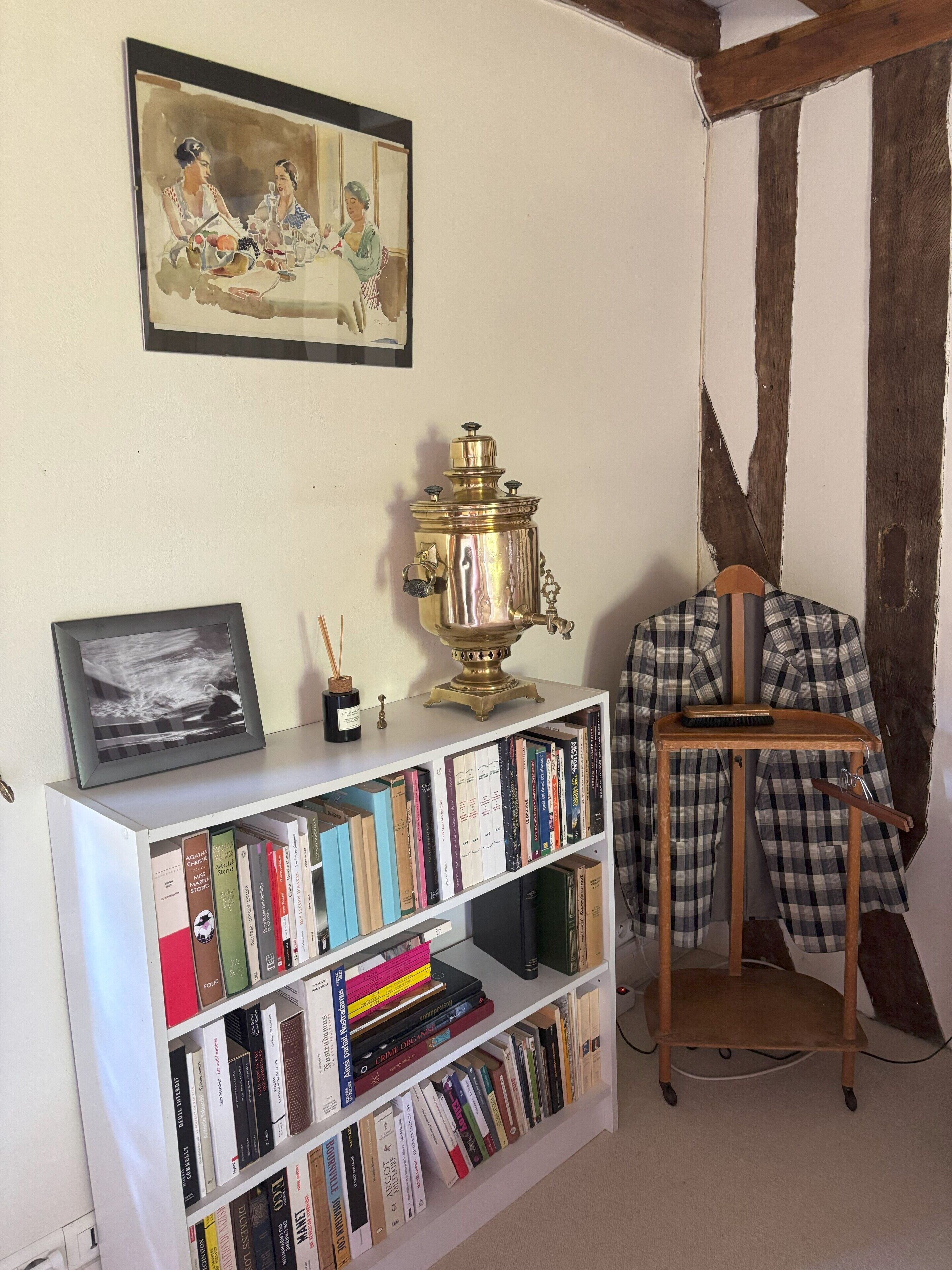 Una habitación con una estantería, un cuadro enmarcado, una estufa de latón y una chaqueta de cuadros sobre una silla.