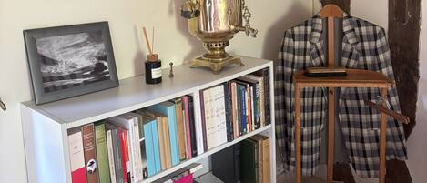 Una habitación con una estantería, un cuadro enmarcado, una estufa de latón y una chaqueta de cuadros sobre una silla.