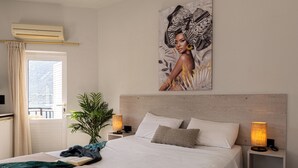 A bedroom with a bed, bedside lamps, a potted plant, a window with curtains, and a painting on the wall. - Bali Seaside Sailor (Mylopotamos)