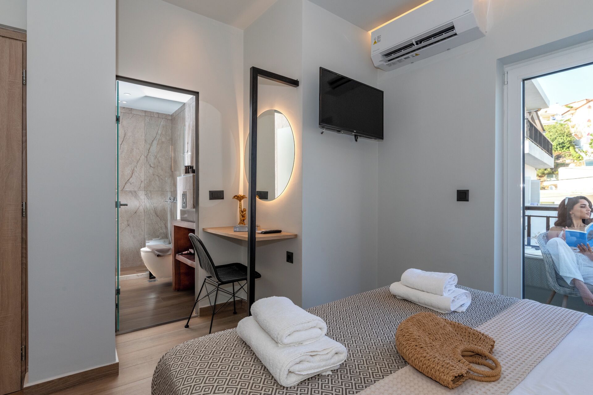 Quarto de hotel moderno com uma cama grande, um espaço de estar com uma poltrona, uma TV fixada na parede e um banheiro visível através de uma porta aberta.