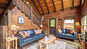 A log cabin interior with a wooden staircase, two sofas, a coffee table, and a television.