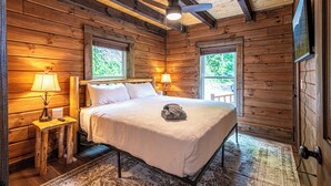 A wooden cabin bedroom with a bed, two windows, a ceiling fan, and a TV on the wall.