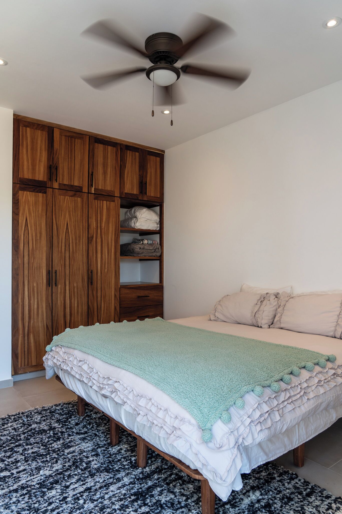 A bedroom with a bed, a ceiling fan, wooden wardrobe, and a black and white rug.