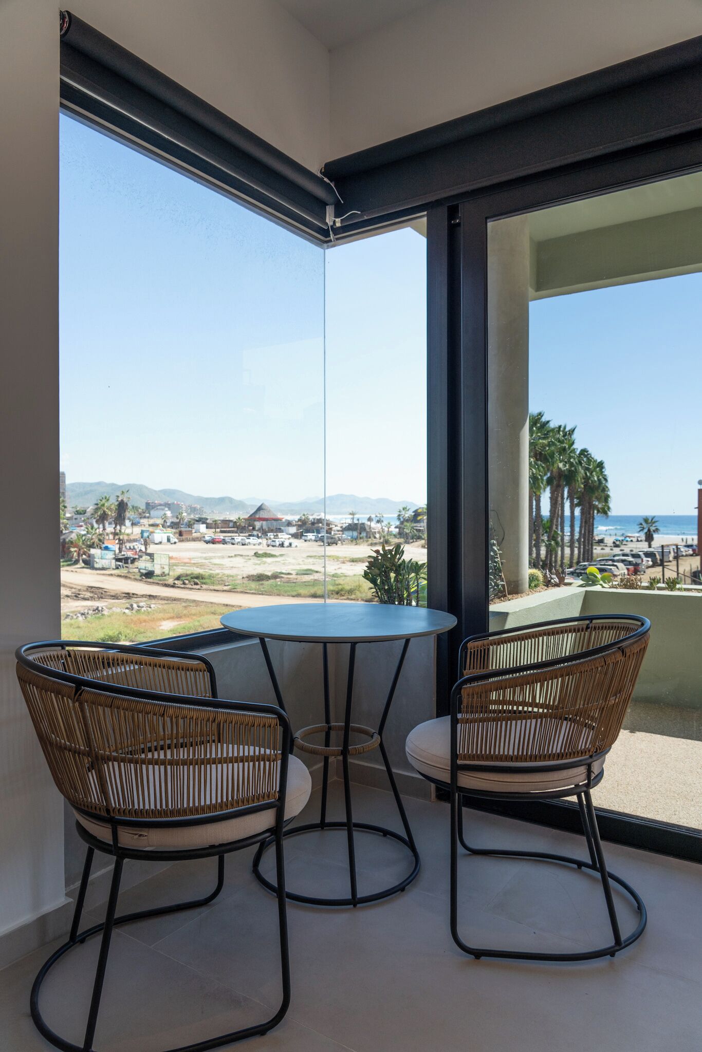 A balcony with a round table and two wicker chairs, overlooking a beach and distant landscape.
