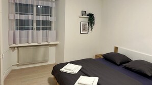 Une chambre à coucher avec un lit, un radiateur, une fenêtre avec des rideaux, une étagère avec une plante et un tableau encadré au mur.