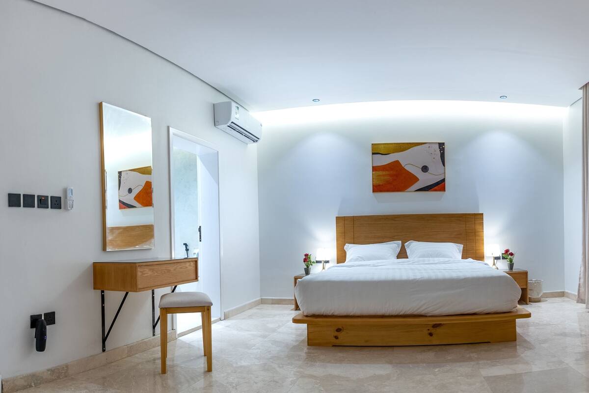 A modern bedroom with a wooden bed, a bedside table, a mirror, a wall-mounted air conditioner, and a wooden shelf.