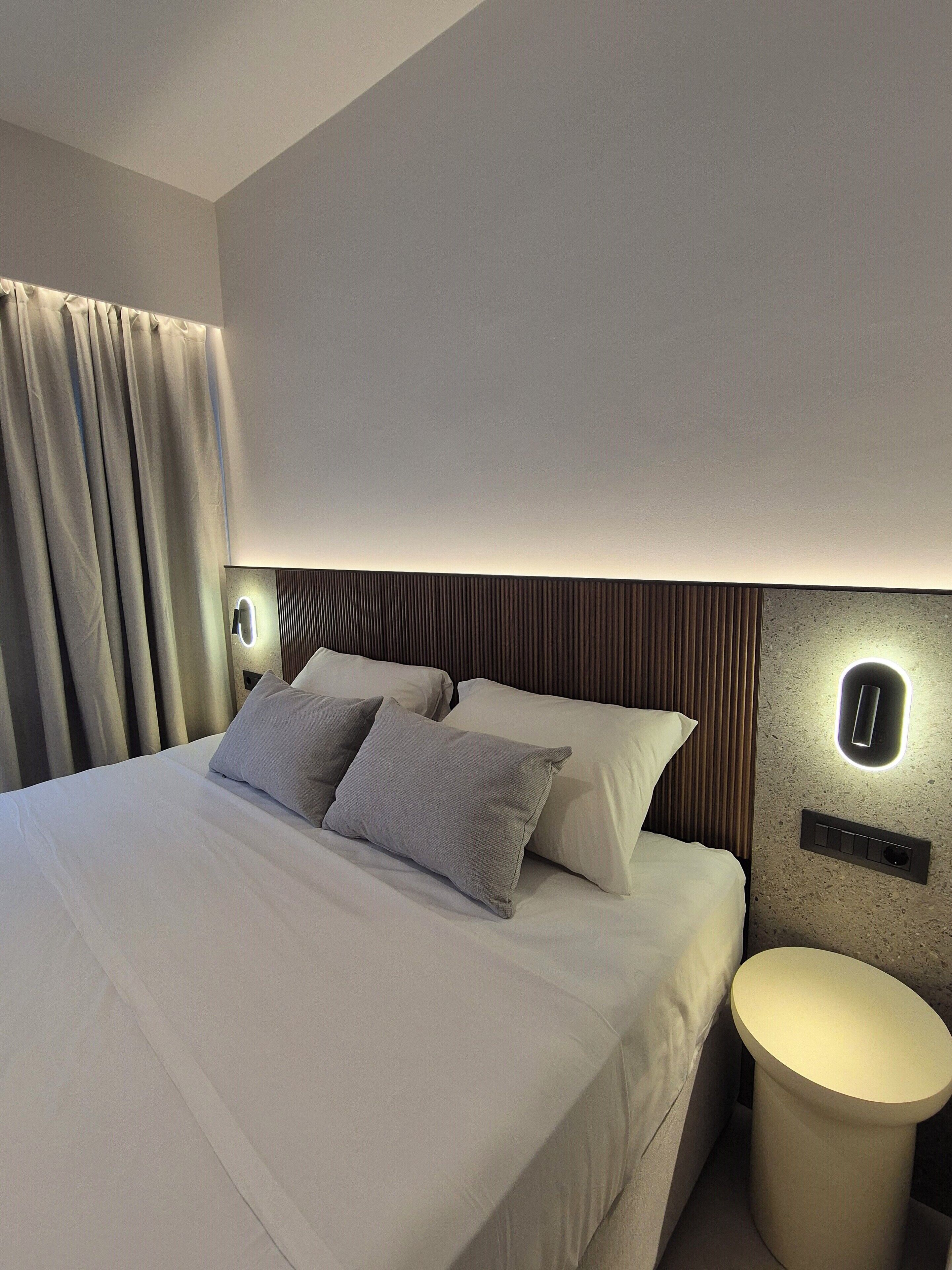 Quarto de hotel moderno com cama bem arrumada, mesa de cabeceira redonda e luminária fixada na parede.