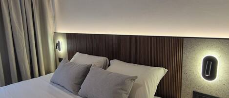 Quarto de hotel moderno com cama bem arrumada, mesa de cabeceira redonda e luminária fixada na parede.