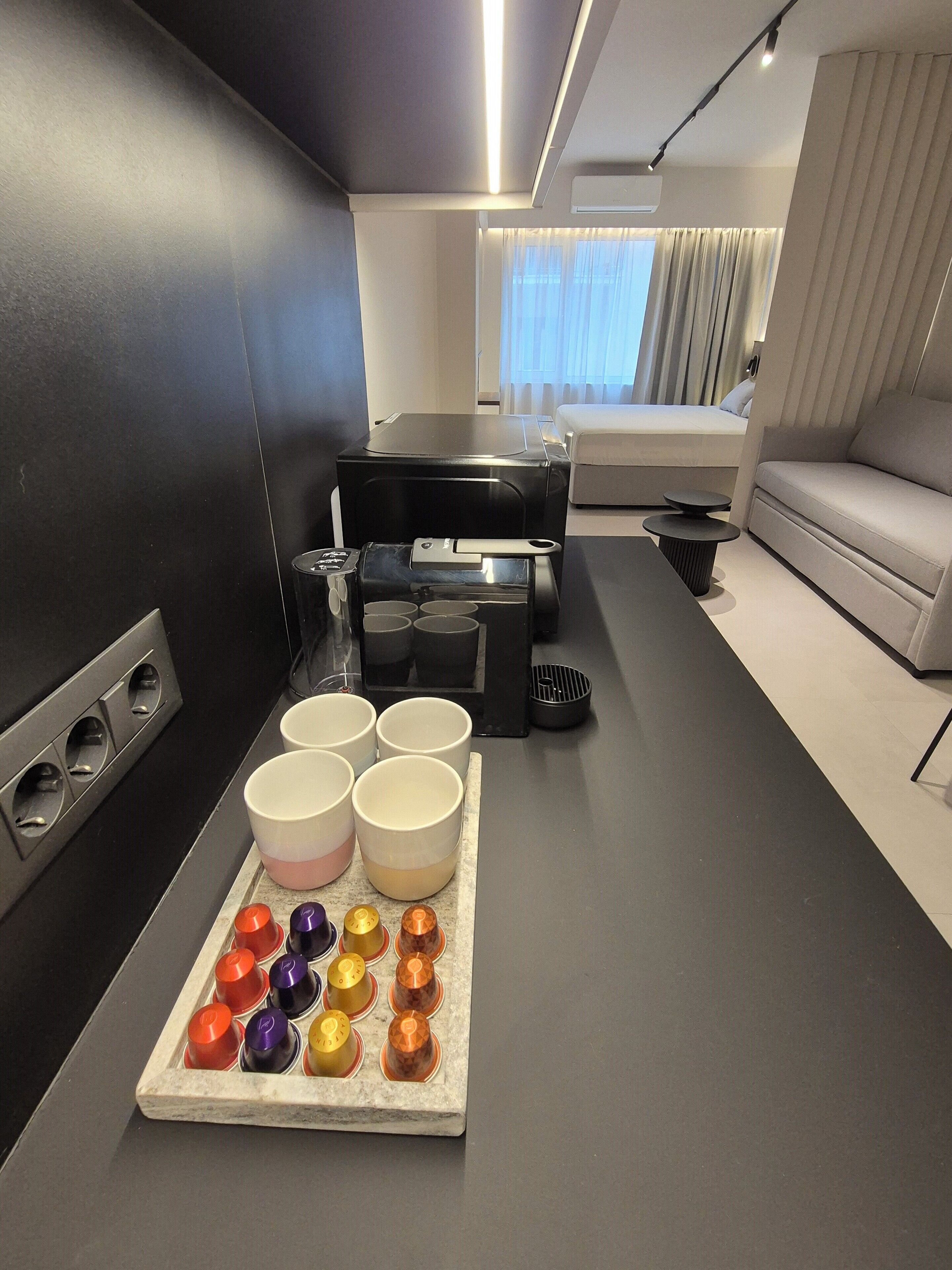 A modern hotel room with a dining area, a sofa, a piano, and a tray of colorful cups.