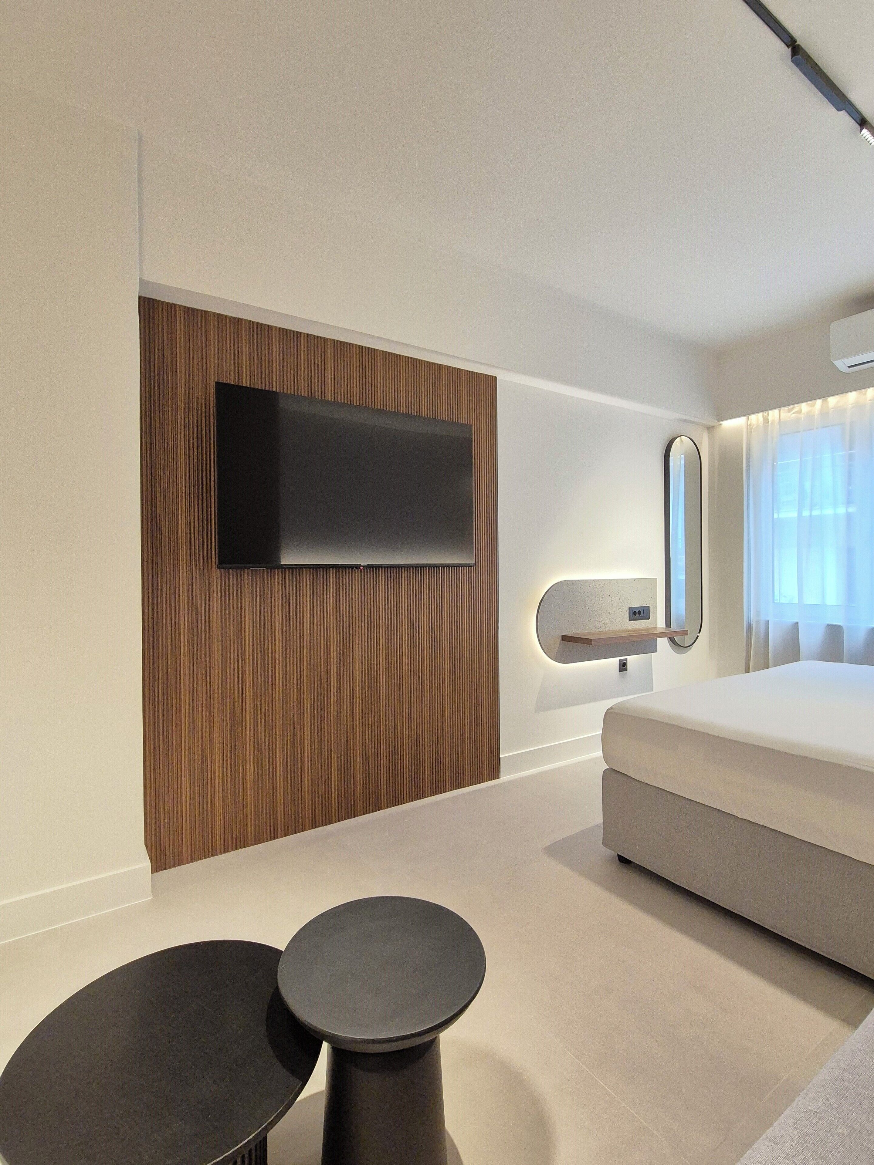 A modern hotel room with a flat-screen TV mounted on a wooden panel wall, a bed with a white mattress, a round black side table, and a round black coffee table.