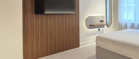 A modern hotel room with a flat-screen TV mounted on a wooden panel wall, a bed with a white mattress, a round black side table, and a round black coffee table.