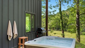 Un jacuzzi al aire libre frente a un edificio con paredes de madera, un taburete de madera y toallas colgadas en ganchos.