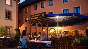 Un espace extérieur aménagé avec des tables et des chaises, une enseigne indiquant « Redstock's Biergarten », et un bâtiment avec de nombreuses fenêtres en arrière-plan.
