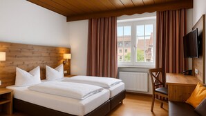 Une chambre d’hôtel avec une tête de lit en bois, deux lits, un bureau, une chaise, un téléviseur et une fenêtre avec des rideaux.