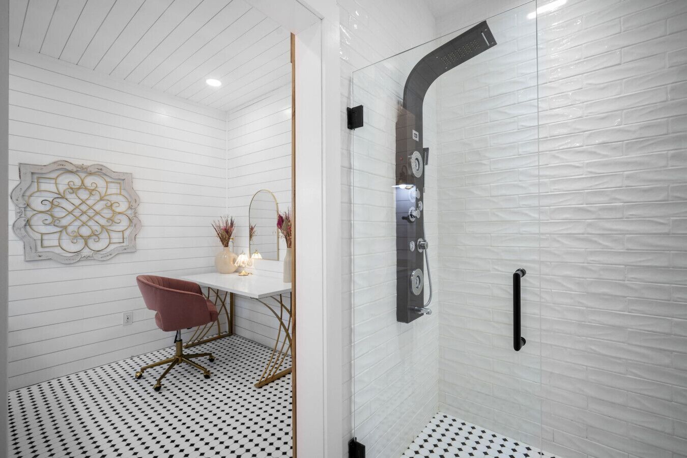 Un baño con pared de azulejos blancos, piso a cuadros blanco y negro, tocador blanco con espejo, una silla rosa y una ducha con cabezal curvo.