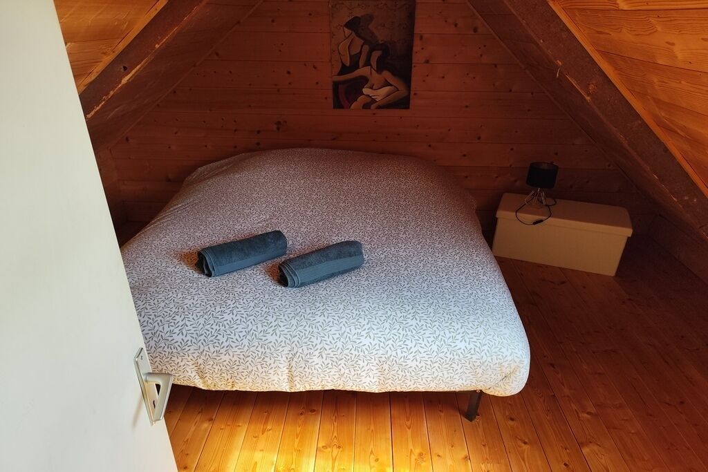 A small, cozy room with a sloped ceiling and wooden walls. There is a bed with a patterned bedspread and two rolled-up blankets. A small white cabinet is against the wall, and a painting hangs above the bed.
