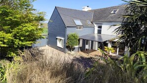 A two-story house with a sloped roof, a small porch, and a garden area with tall grasses and trees. - Charming family home (Taulé)