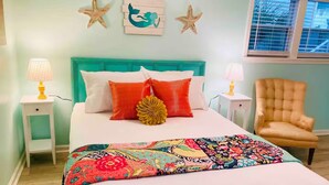 Une chambre avec une tête de lit turquoise, du linge de lit blanc et une couverture colorée. On peut voir des décorations en forme d’étoiles sur le mur, une fenêtre avec des stores et une chaise près de la fenêtre.
