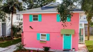 Une maison à deux étages, avec une façade rose et des volets verts, un porche à l’avant et un petit jardin agrémenté de plantes et de fleurs.