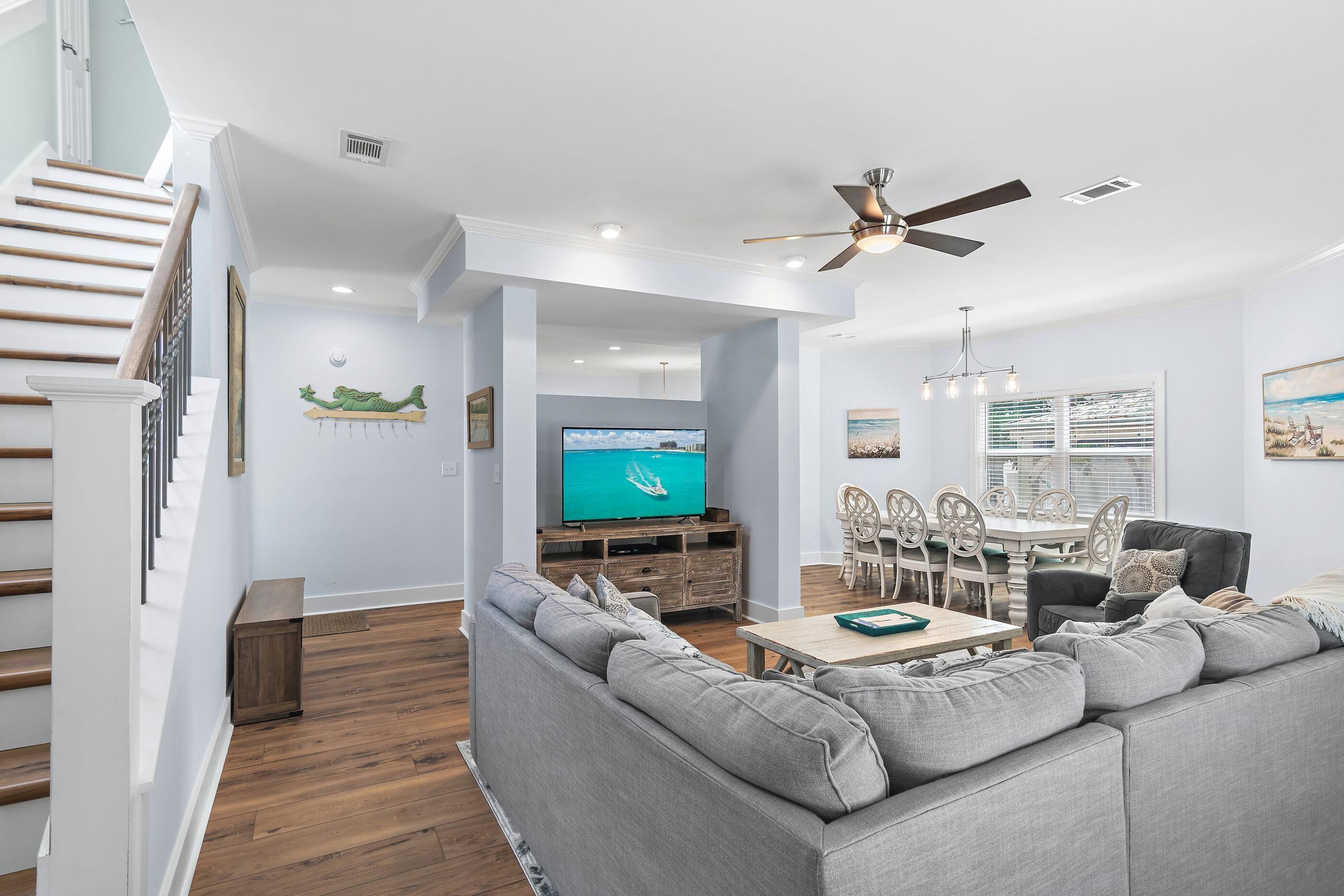 A modern living room with a grey sofa, wooden flooring, a TV, a dining area with a table and chairs, and a ceiling fan.