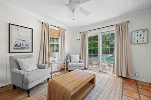 A spacious living room with a grey sofa, a wooden bench, and a white armchair. There is a ceiling fan, a large window with curtains, and a framed picture on the wall. - "Palm Beach Paradise: Cozy 1BR + Pool, Minutes to Everything" (West Palm Beach)