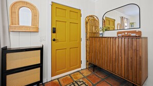 Un pasillo con una puerta amarilla, un espejo, un banco de mimbre y un armario de madera.