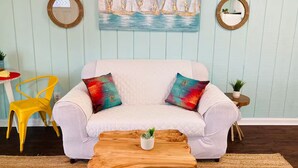 Un salón con un sofá blanco, cojines coloridos, una mesa de centro de madera con una planta, un cuadro de veleros y dos espejos redondos en la pared.