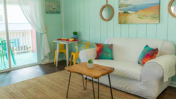 Una habitación con un sofá blanco, almohadones coloridos, una mesita de centro de madera, una pequeña mesa con sillas y un cuadro en la pared.